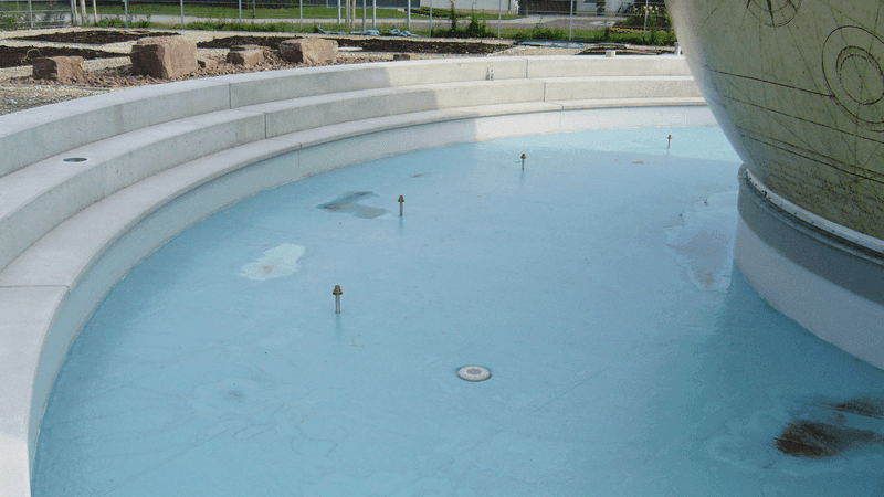 Wasserbecken einer Außenanlage mit heller Terrassenstruktur und dekorativer Weltkugel, Teil eines gestalteten Freiraumbereichs.