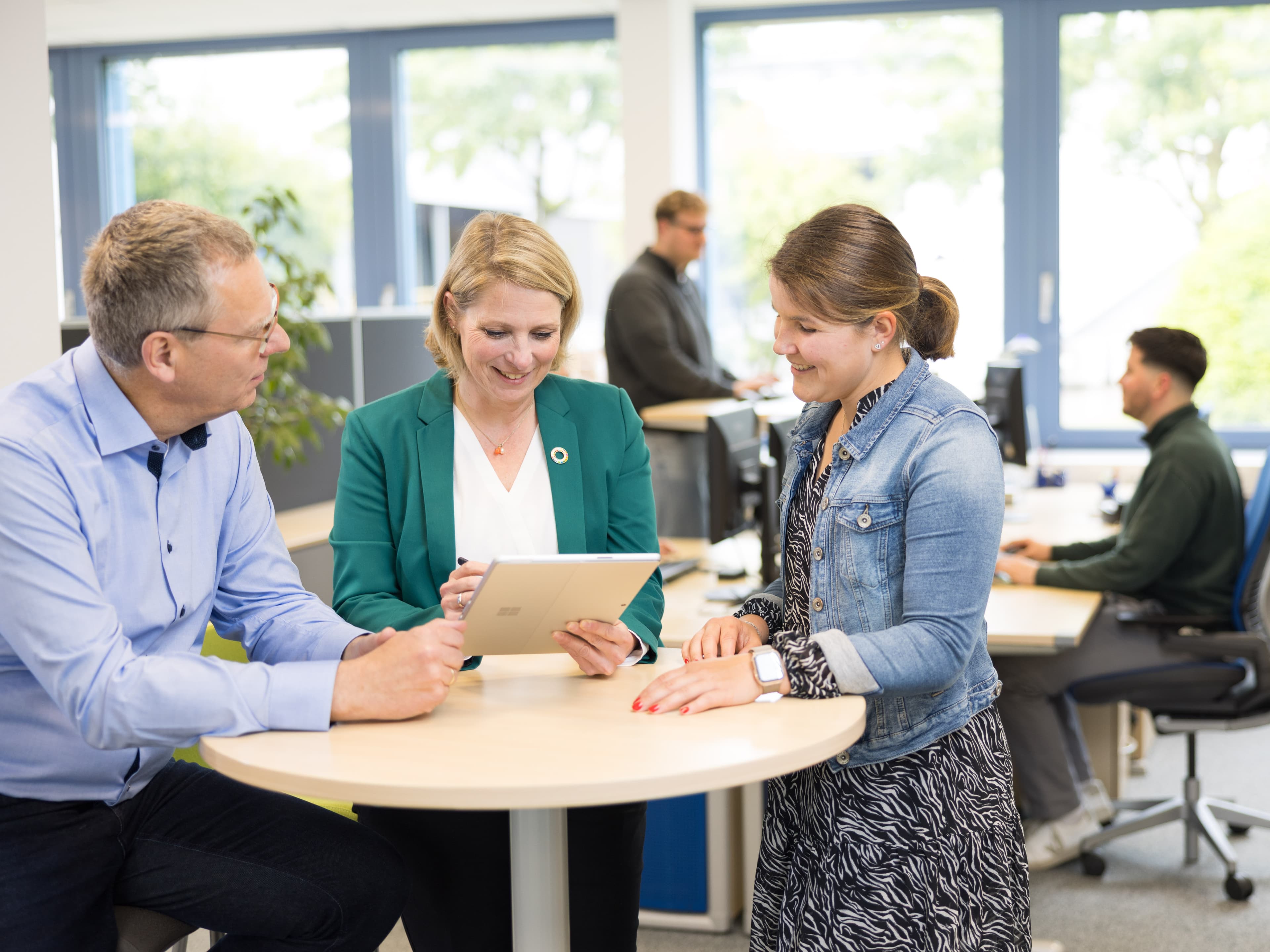 Fachkräfte besprechen Projektunterlagen an einem Besprechungstisch in einem modernen Büro mit Teamarbeit im Hintergrund.