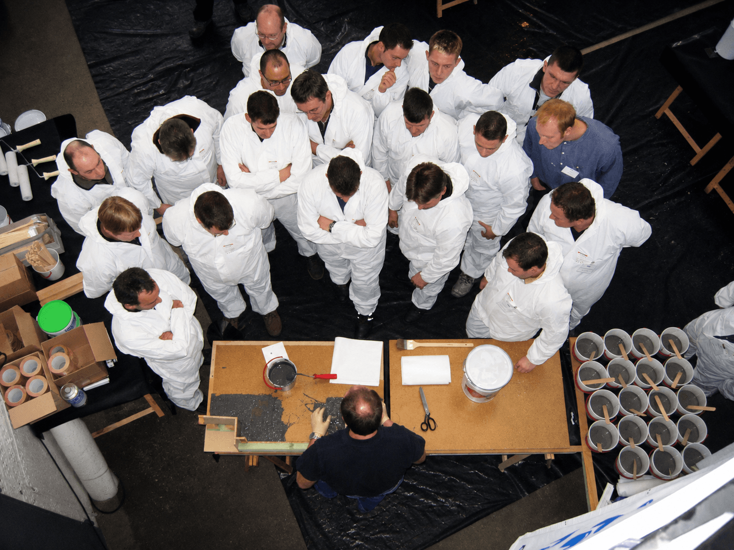 Teilnehmer stehen im Praxisseminar um ein Modell und verfolgen eine praktische Demonstration zur Verarbeitung von Triflex Abdichtungs- und Beschichtungssystemen.