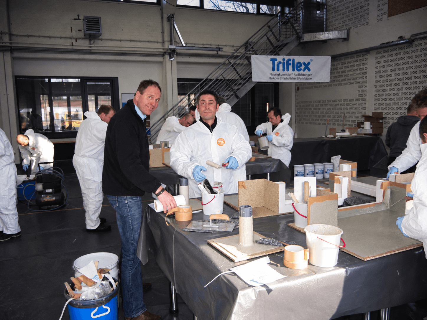 Teilnehmer und Triflex Anwendungstechniker stehen im Praxisseminar an einem Arbeitstisch mit Modellen, Werkzeugen und Materialien.
