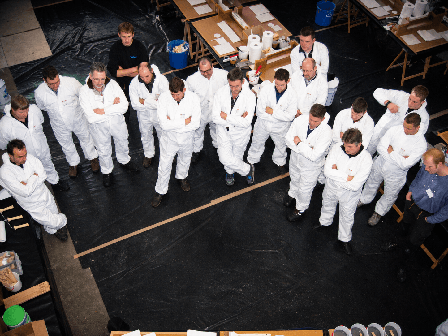 Teilnehmer stehen im Praxisseminar in Schutzanzügen um ein Modell und verfolgen eine praktische Vorführung zur Verarbeitung von Triflex Systemen.