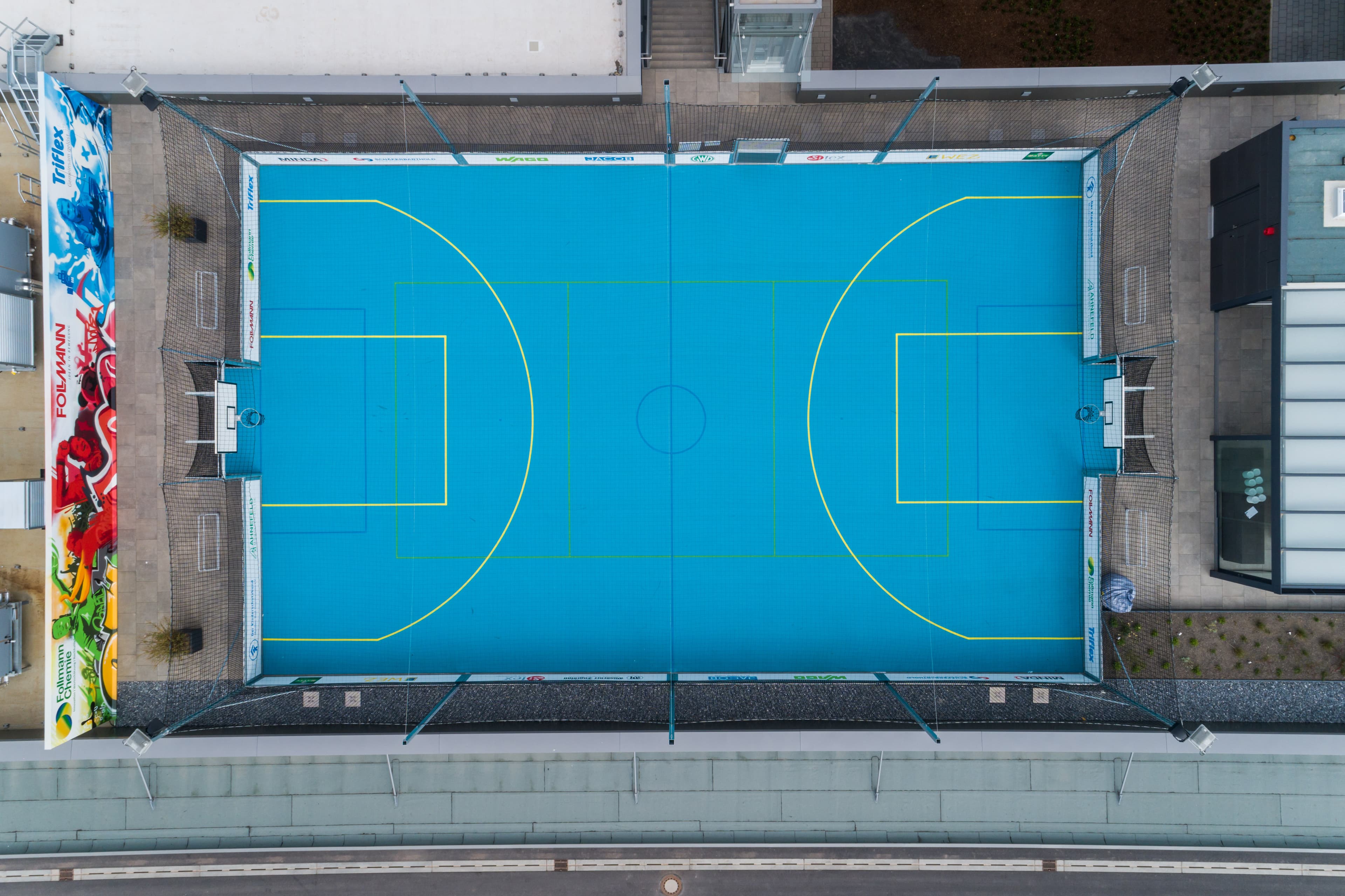 A blue-coated sports field on a building roof with court markings in an urban setting.