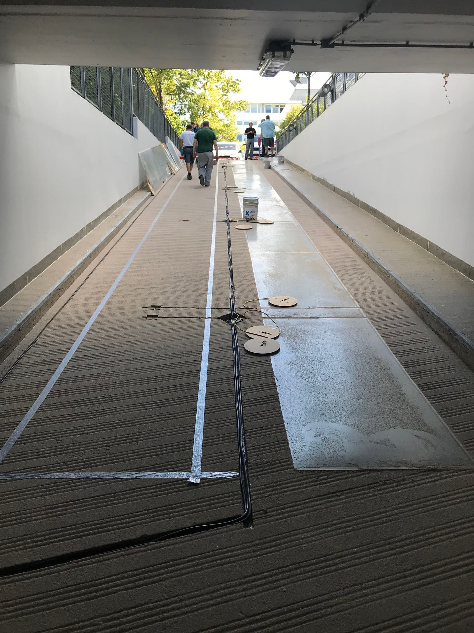Sanierung einer Tiefgaragenzufahrt mit neuem Bodenbelag und markierten Führungsschienen; Handwerker arbeiten am oberen Rampenbereich.