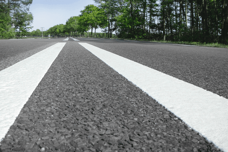 Nahaufnahme frisch aufgebrachter weißer Straßenmarkierungen auf Asphaltfahrbahn in grüner Umgebung.