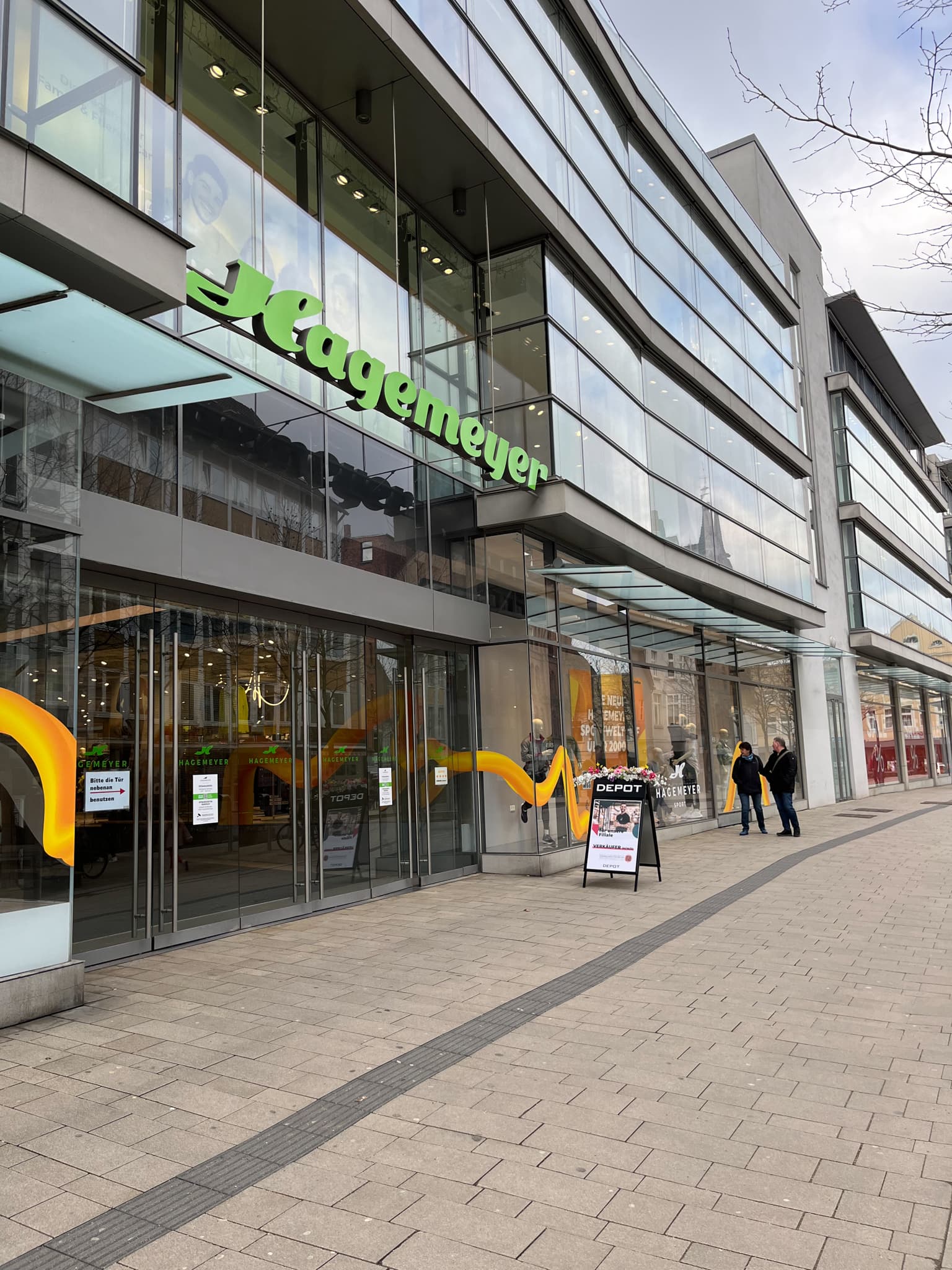 Moderne Glasfassade des Hagemeyer-Gebäudes in einer Einkaufsstraße in Minden, mit gut sichtbarem Eingangsbereich und Passanten vor dem Geschäft.