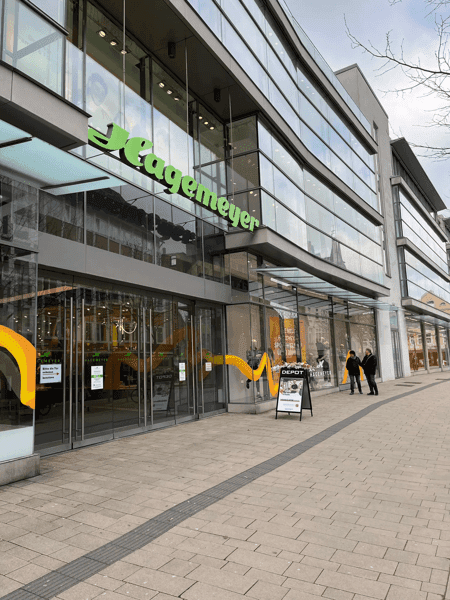 Moderne Glasfassade des Hagemeyer-Gebäudes in einer Einkaufsstraße in Minden, mit gut sichtbarem Eingangsbereich und Passanten vor dem Geschäft.