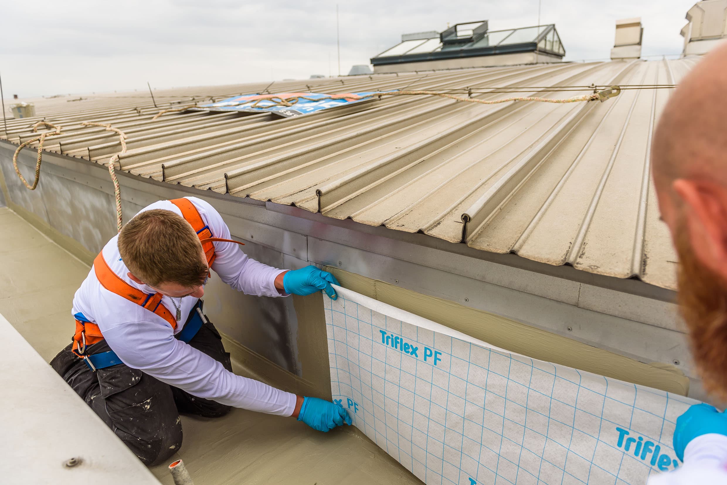 Contractors are installing a waterproofing membrane along the edge where a sheet-metal roof meets the roof structure.