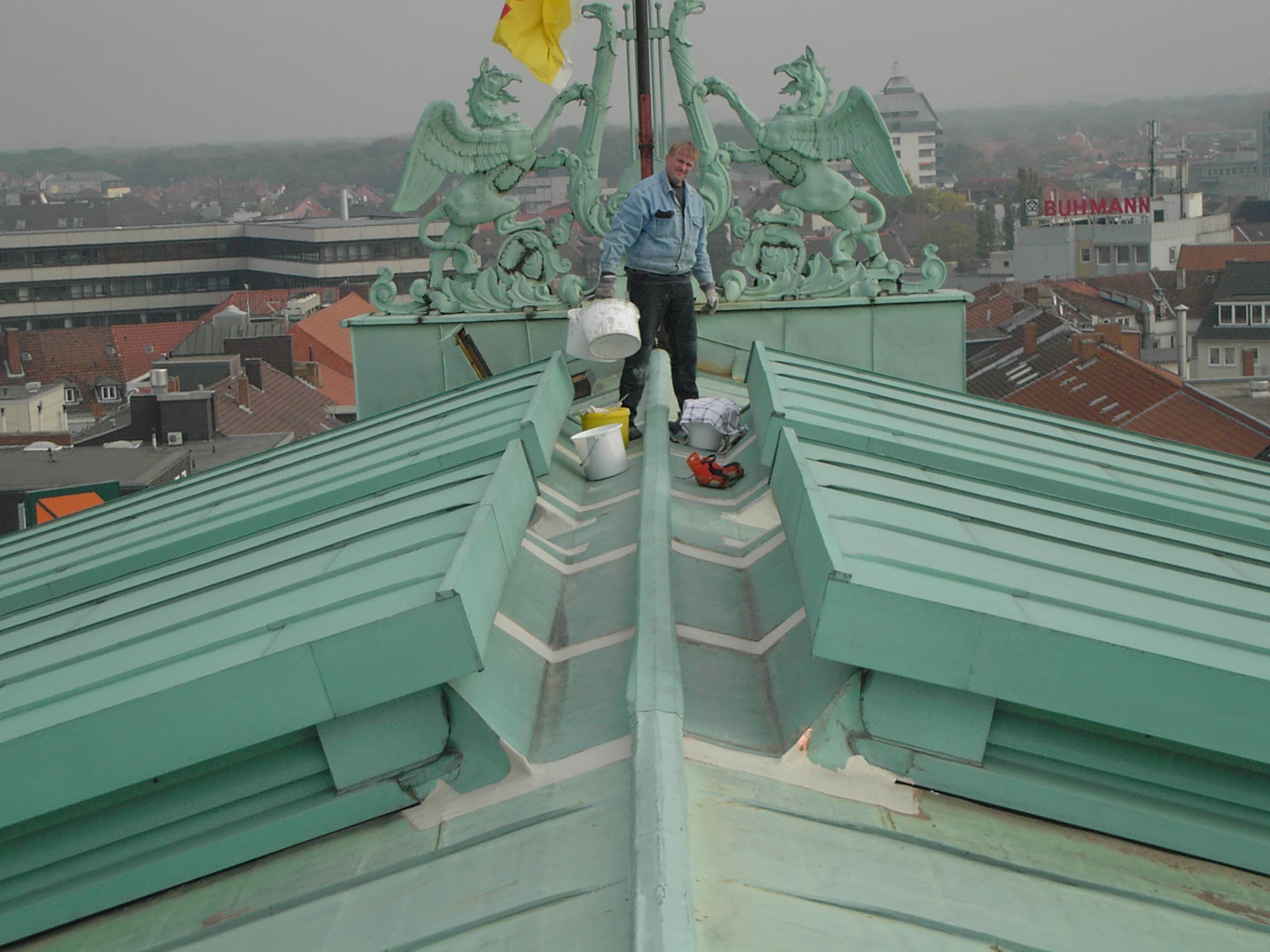 Handwerker bei Wartungsarbeiten auf einem großflächigen Kupferdach mit grüner Patina, Stadtansicht im Hintergrund.