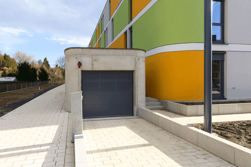 Zufahrt zu einer Tiefgarage mit modernem Rolltor und farbig gestalteter Gebäudefassade nach Sanierung.