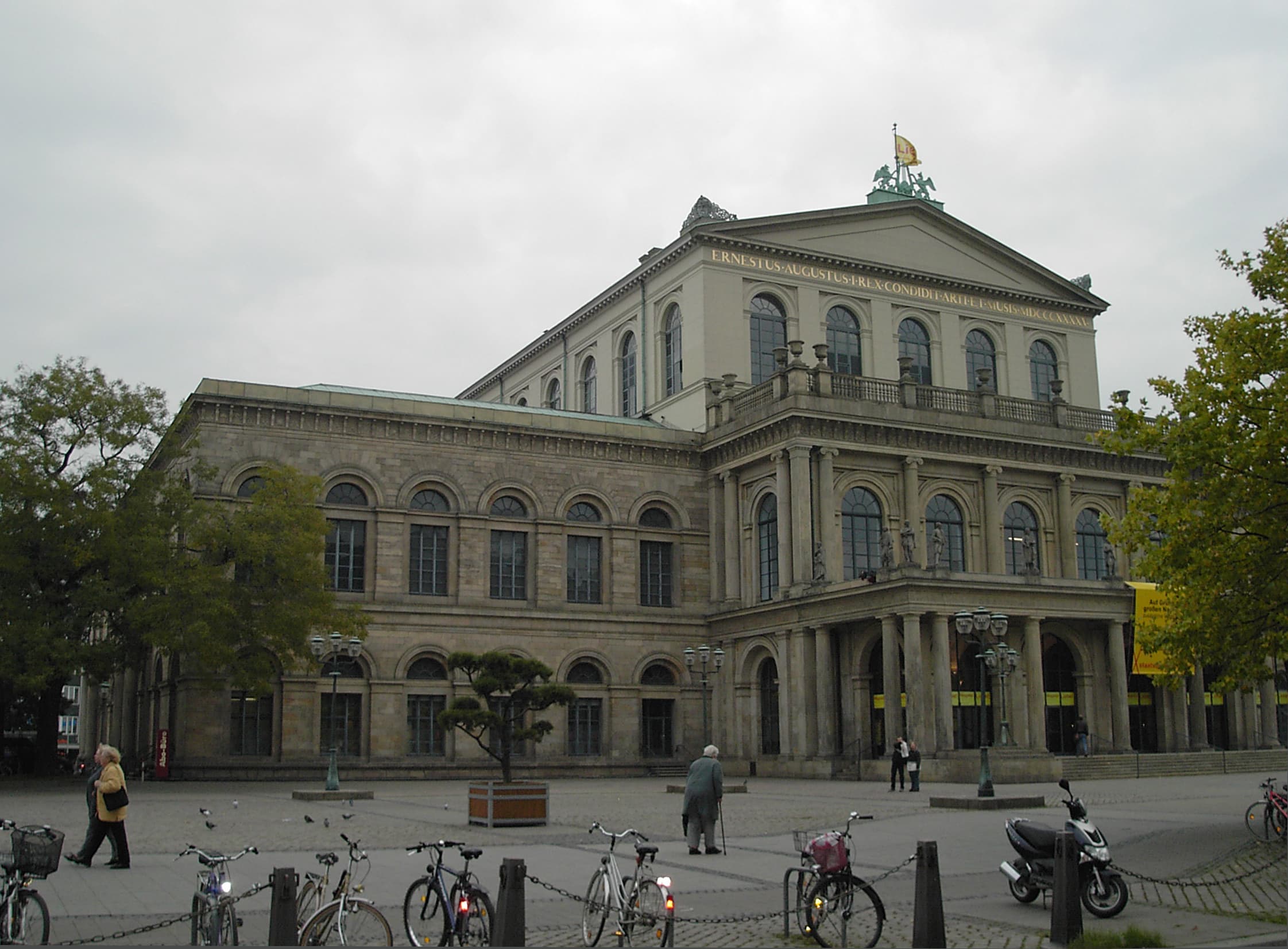 Historisches Opernhaus mit klassizistischer Fassade an einem städtischen Platz, Fahrräder und Passanten im Vordergrund.