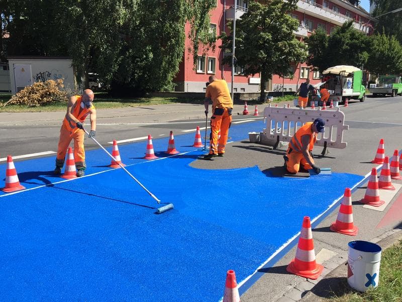 Straßenbau-Team trägt blaue Markierungsbeschichtung auf eine abgesperrte Fahrbahn auf.