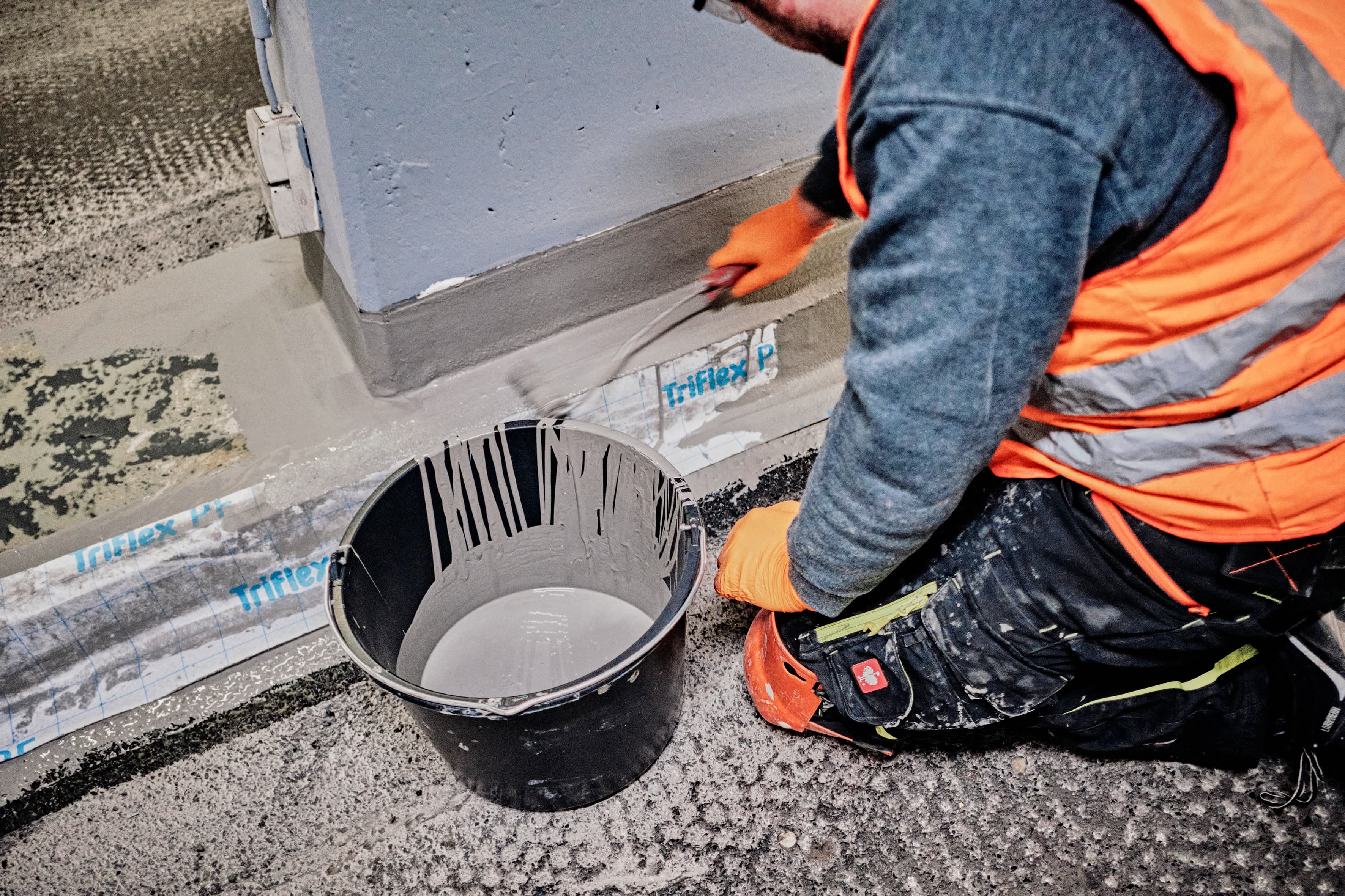 Handwerker trägt eine flüssige Abdichtungsmasse an einer Gebäudekante auf; Verarbeitung von Bauwerksabdichtung mit Pinsel neben einem Eimer Beschichtungsmaterial.