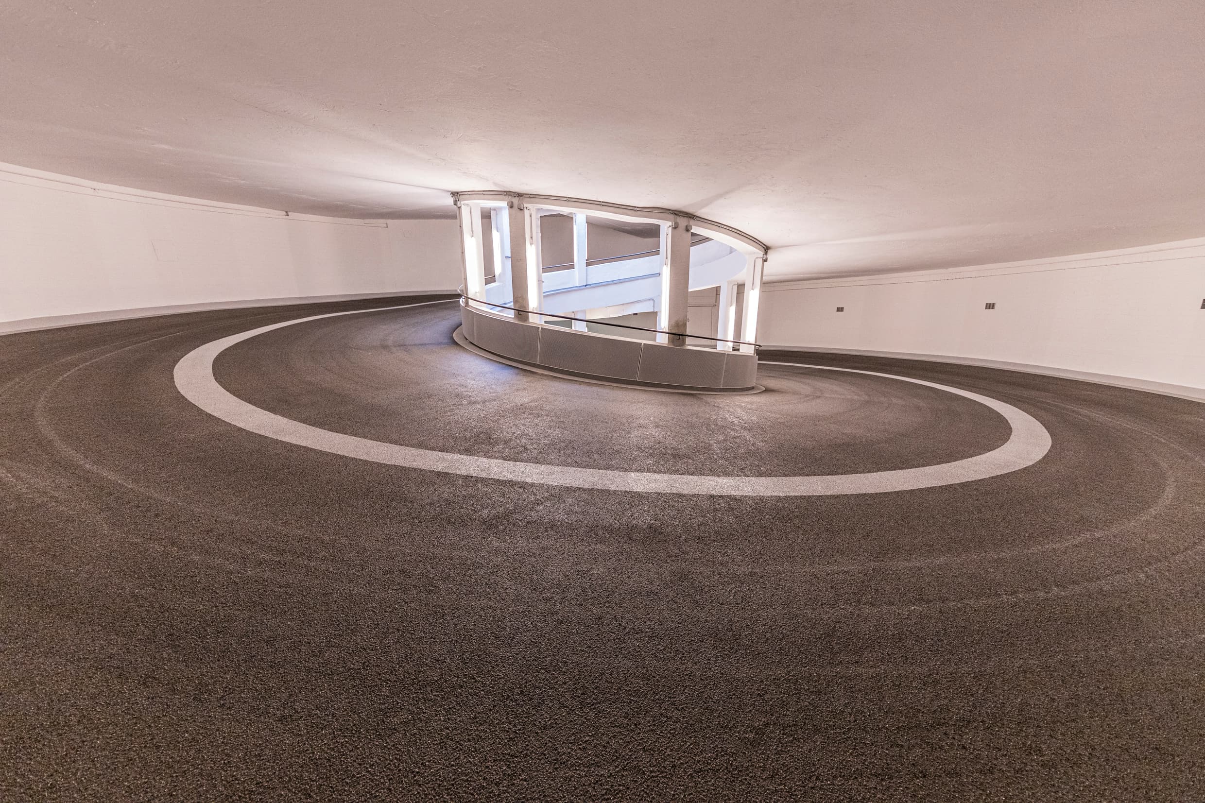 Rund verlaufende Parkhausrampe mit markierten Fahrspuren und beschichteter Oberfläche in einem hellen Innenbereich, fotografisch dargestellt.