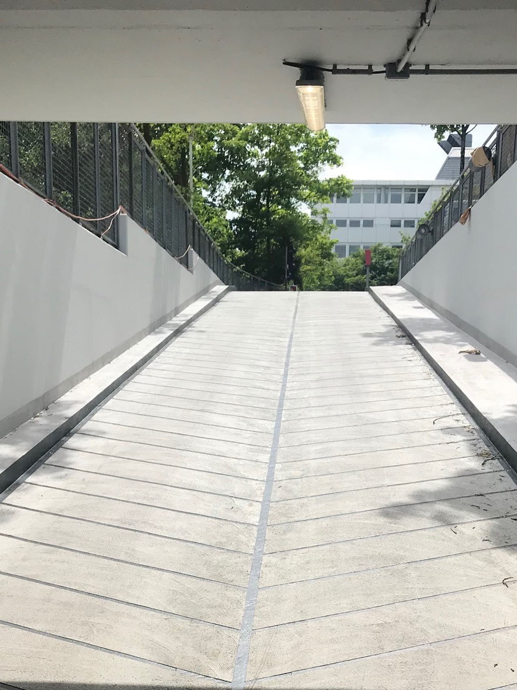 Betonrampe in einer modernen Tiefgaragenzufahrt mit weißen Seitenwänden und Geländern, im Hintergrund grüne Bäume und ein Bürogebäude bei Tageslicht.